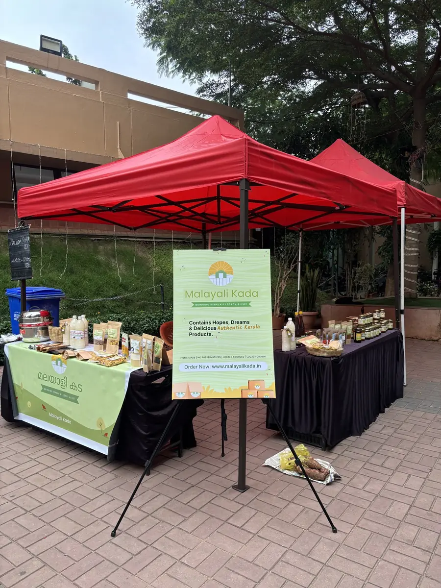 A Malayali Kada stall under a red canopy at a society event, with a standing sign beside the table