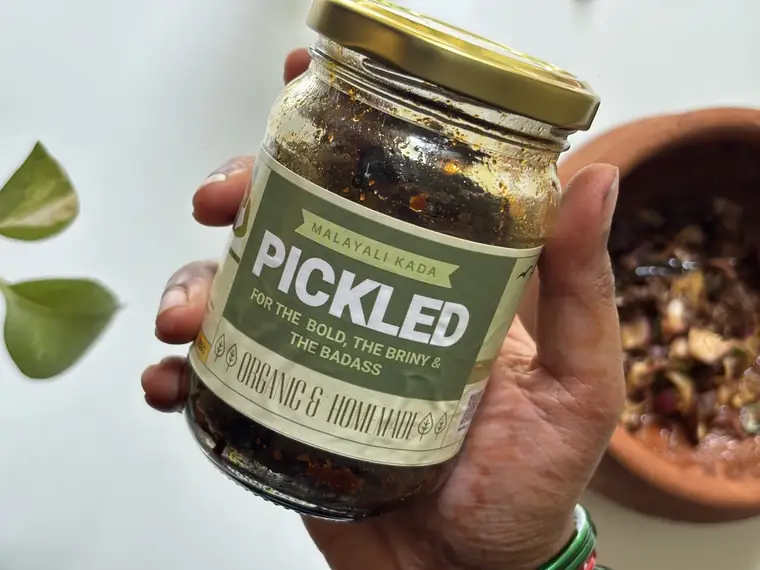 A hand holding a jar of homemade pickle with a Malayali Kada label