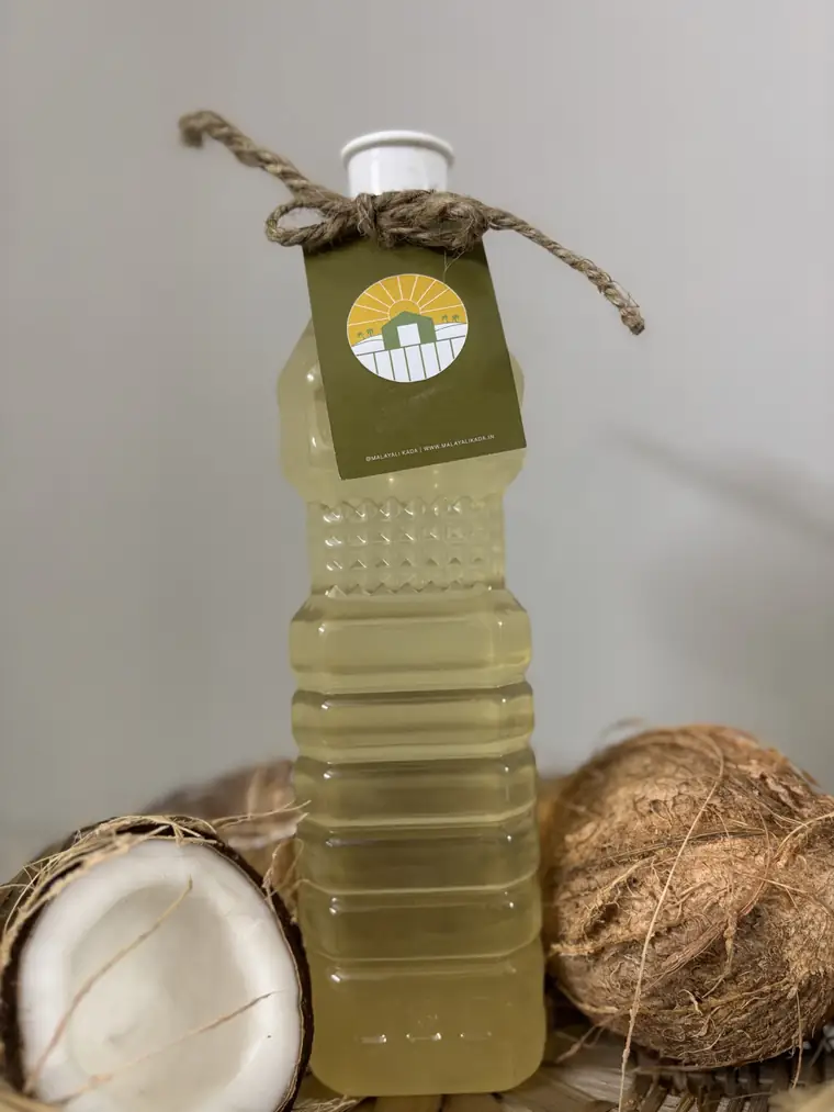 Cold pressed coconut oil bottle with a round Malayali Kada tag, beside coconut halves